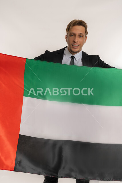 Union Day 1971, Getting to know the cultures and history of the Emirates, Portrait of a smiling foreign businessman wearing a formal suit holding a flag to celebrate the UAE National Day, December 2, standing straight and firm with expressions of pride and honor, looking at the camera with gestures of pleasure, white background