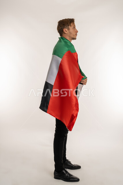 الوقوف باستقامة وثبات بتعابير الفخر والسرور، ذكرى تأسيس الاتحاد 1971، التعرف على ثقافات وتاريخ الامارات، بورتريه من الجانب لرجل اعمال اجنبي يرتدي البدلة الرسمية يحمل العلم على ظهره للاحتفال باليوم بالوطني الاماراتي 2 ديسمبر، الطول الكامل للجسم، خلفية بيضاء