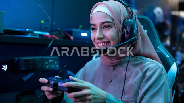 Modern entertaining technological techniques, fun and entertainment with video games, spending free time playing entertaining electronic games, a smiling veiled Saudi Arabian Gulf young woman wearing headphones and holding a joystick with gestures of integration and focus