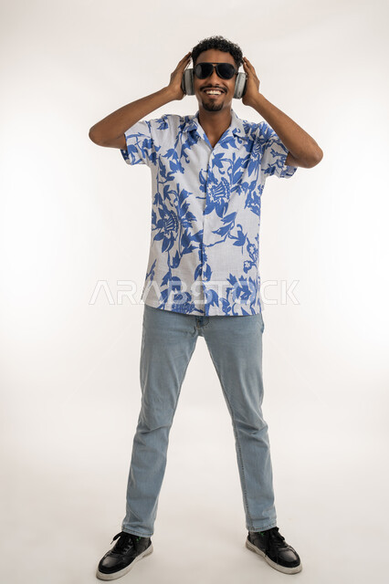 مفهوم الغناء والطرب، رجل سعودي يستخدم تقنية الهيدفون، بورتريه لشاب اسمر عربي خليجي اماراتي يرتدي الزي الكاجوال والنظارة الشمسية يضع سماعة الراس يستمع الى الاغاني بتعابير الاندماج والسرور، قضاء اوقات ممتعة بالاستماع الى الموسيقى، صورة الجسم بالكامل، خلفية بيضاء