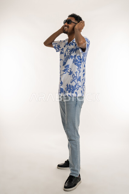 دمج التسلية بالتقنية الحديثة، رجل سعودي يضع يده فوق الهيدفون، مفهوم الغناء والطرب والاندماج، بورتريه لشاب اسمر عربي خليجي اماراتي يرتدي الزي الكاجوال والنظارة الشمسية وسماعة الرأس يستمع الى الانغام الموسيقية بتعابير السرور، صورة الجسم بالكامل، خلفية بيضاء