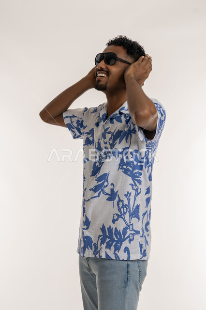 قضاء اوقات ممتعة بالاستماع الى الموسيقى، رجل سعودي يضع يده فوق الهيدفون الحديثة، مفهوم الغناء والطرب، بورتريه لشاب اسمر عربي خليجي اماراتي يرتدي الزي الكاجوال والنظارة الشمسية يضع سماعة الرأس ويستمع الى الانغام الموسيقية بتعابير الاندماج والسرور، خلفية بيضاء