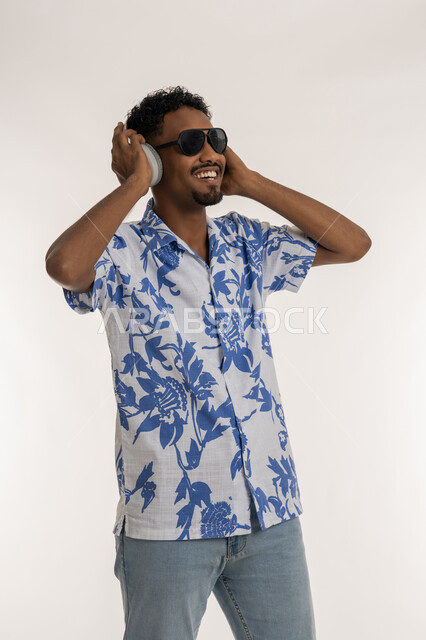 Having a good time listening to music, a Saudi man putting his hand on the modern headphone, the concept of singing and music, a portrait of a dark-skinned young Arab Gulf Emirati man wearing casual clothes and sunglasses, putting on headphones and listening to musical tunes with expressions of integration and pleasure, white background