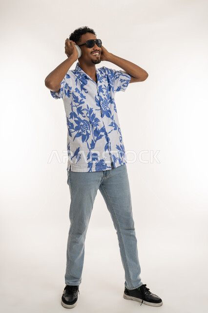 دمج التسلية بالتقنية الحديثة، رجل سعودي يضع يده فوق الهيدفون، مفهوم الغناء والطرب والاندماج، بورتريه لشاب اسمر عربي خليجي اماراتي يرتدي الزي الكاجوال والنظارة الشمسية وسماعة الرأس يستمع الى الانغام الموسيقية بتعابير السرور، صورة الجسم بالكامل، خلفية بيضاء