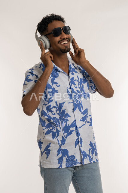 قضاء اوقات ممتعة بالاستماع الى الموسيقى، رجل سعودي يضع يده فوق الهيدفون الحديثة، مفهوم الغناء والطرب، بورتريه لشاب اسمر عربي خليجي اماراتي يرتدي الزي الكاجوال والنظارة الشمسية يضع سماعة الرأس ويستمع الى الانغام الموسيقية بتعابير الاندماج والسرور، خلفية بيضاء