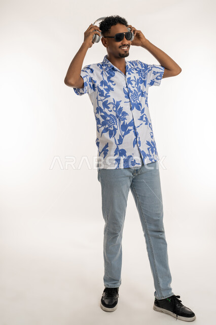 مفهوم الغناء والطرب، قضاء اوقات ممتعة بالاستماع الى الموسيقى، رجل سعودي يستخدم تقنية الهيدفون الحديثة، بورتريه لشاب اسمر عربي خليجي اماراتي مبتسم يرتدي الزي الكاجوال والنظارة الشمسية يمسك بيده سماعة الرأس بتعابير الفرح والسرور، صورة الجسم بالكامل، خلفية بيضاء