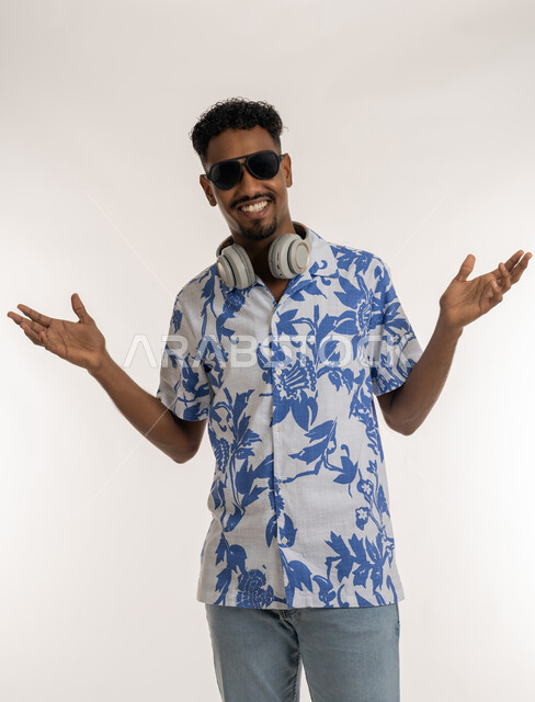 الرقص والتفاعل والاندماج مع الانغام الموسيقية، النظر الى الكاميرا بتعابير السرور والسعادة، رجل سعودي يستخدام تقنية الهيدفون الحديثة، بورتريه لشاب اسمر عربي خليجي اماراتي يرتدي الزي الكاجوال والنظارة الشمسية يضع سماعة الرأس حول عنقه، خلفية بيضاء