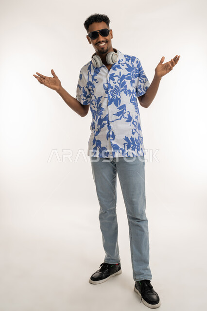 النظر الى الكاميرا بتعابير السرور والسعادة، رجل سعودي يستخدام تقنية الهيدفون، بورتريه لشاب اسمر عربي خليجي اماراتي يرتدي الزي الكاجوال والنظارة الشمسية يضع سماعة الرأس حول عنقه، الرقص والتفاعل والاندماج مع الانغام الموسيقية، صورة الجسم بالكامل، خلفية بيضاء