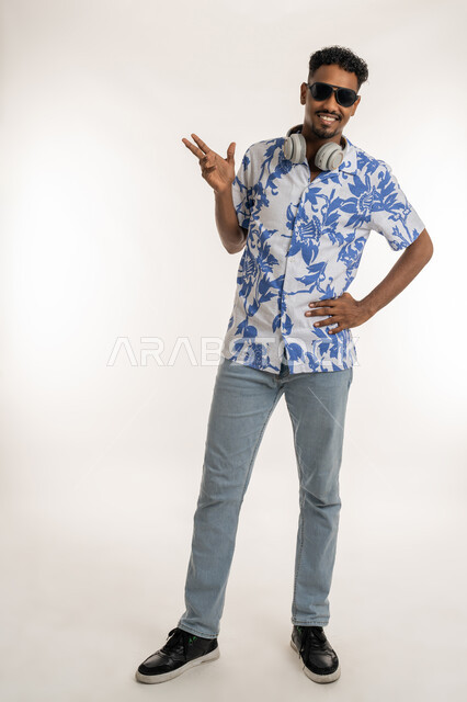 النظر الى الكاميرا بتعابير السرور والسعادة، رجل سعودي يستخدام تقنية الهيدفون، بورتريه لشاب اسمر عربي خليجي اماراتي يرتدي الزي الكاجوال والنظارة الشمسية يضع سماعة الرأس حول عنقه، الرقص والتفاعل والاندماج مع الانغام الموسيقية، صورة الجسم بالكامل، خلفية بيضاء