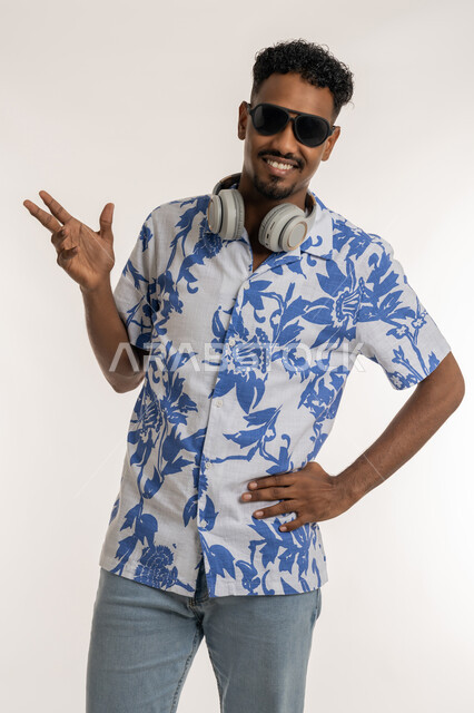 Dancing, interacting and merging with the musical tunes, looking at the camera with expressions of pleasure and happiness, a Saudi man using modern headphone technology, a portrait of a dark-skinned young Arab Gulf Emirati man wearing casual clothes and sunglasses, wearing a headphone around his neck, white background