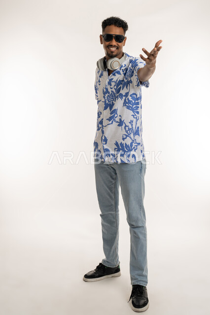 الرقص والتفاعل والاندماج مع الانغام الموسيقية، النظر الى الكاميرا بتعابير السرور، رجل سعودي يستخدام تقنية الهيدفون، بورتريه لشاب اسمر عربي خليجي اماراتي يرتدي الزي الكاجوال والنظارة الشمسية يضع سماعة الرأس حول عنقه، صورة الجسم بالكامل، خلفية بيضاء