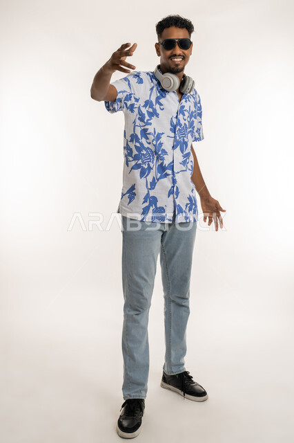 الرقص والتفاعل والاندماج مع الانغام الموسيقية، النظر الى الكاميرا بتعابير السرور، رجل سعودي يستخدام تقنية الهيدفون، بورتريه لشاب اسمر عربي خليجي اماراتي يرتدي الزي الكاجوال والنظارة الشمسية يضع سماعة الرأس حول عنقه، صورة الجسم بالكامل، خلفية بيضاء