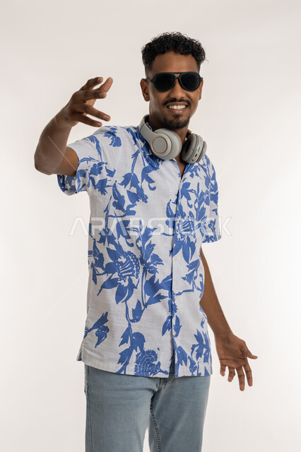 قضاء اوقات ممتعة بالاستماع الى الموسيقى، الرقص والتفاعل مع الانغام الموسيقية، النظر الى الكاميرا بتعابير السرور، رجل سعودي يستخدام تقنية الهيدفون، بورتريه لشاب اسمر عربي خليجي اماراتي يرتدي الزي الكاجوال والنظارة الشمسية يضع سماعة الرأس حول عنقه، خلفية بيضاء