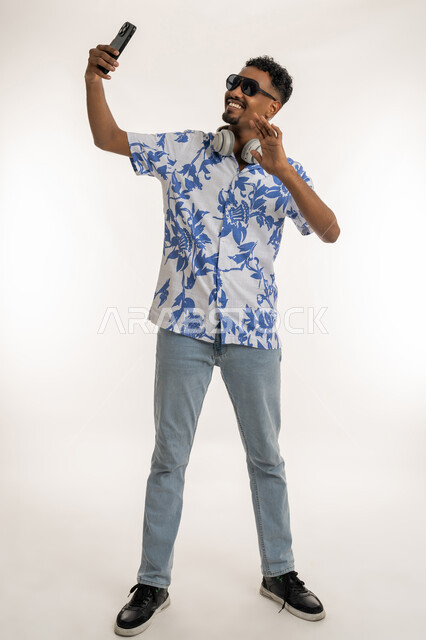 إلقاء التحية والترحيب والسلام، بورتريه لشاب اسمر عربي خليجي اماراتي يرتدي الزي الكاجوال والنظارات الشمسية يقوم باجراء مكالمة فيديو باستخدام الهاتف المحمول، رجل سعودي يتواصل مع الاهل والاصدقاء عبر الانترنت، صورة الجسم بالكامل، خلفية بيضاء