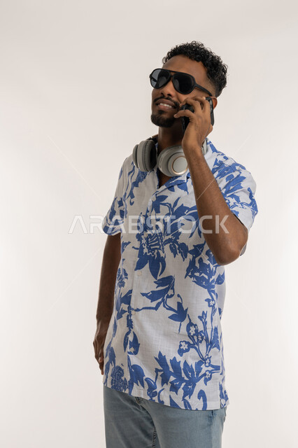 مفهوم الاناقة والشياكة، رجل سعودي يستخدم الاجهزة الحديثة، بورتريه لشاب اسمر عربي خليجي اماراتي مبتسم يرتدي الزي الكاجوال والنظارات الشمسية يضع سماعة الرأس حول عنقه يقوم بإجراء مكالمة هاتفية باستخدام الجوال، التواصل مع الاهل والاصدقاء، خلفية بيضاء