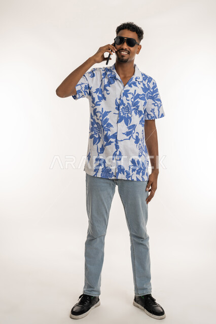 سماع اخبار سعيدة، رجل سعودي يقوم باستخدام الاجهزة الحديثة، بورتريه لشاب اسمر عربي خليجي اماراتي مبتسم يرتدي الزي الكاجوال والنظارات الشمسية يقوم بإجراء مكالمة هاتفية باستخدام الهاتف المحمول، التواصل مع الاهل والاصدقاء، صورة الجسم بالكامل، خلفية بيضاء