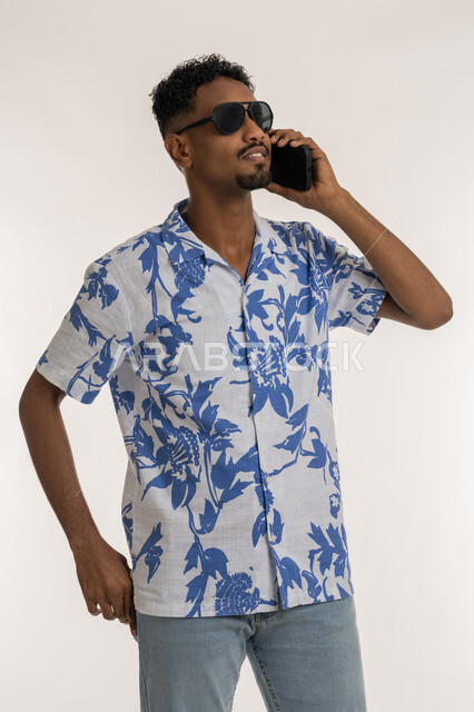 مفهوم الاناقة والشياكة، رجل سعودي يقوم باستخدام الاجهزة الحديثة في الحياة اليومية، بورتريه لشاب اسمر عربي خليجي اماراتي يرتدي الزي الكاجوال والنظارات الشمسية يقوم بإجراء مكالمة هاتفية باستخدام الجوال، التواصل مع الاهل والاصدقاء، خلفية بيضاء