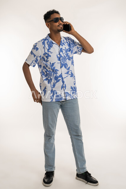 Communication with family and friends, the concept of elegance and chic, a Saudi man using modern devices in daily life, a portrait of a dark-skinned young Arab Gulf Emirati man wearing casual clothes and sunglasses making a phone call using a mobile phone, a full body image, white background