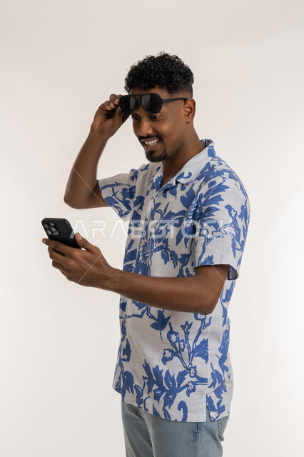 Viewing happy news, a Saudi man using modern devices, a portrait of a smiling dark-skinned young Arab Gulf Emirati man wearing casual clothes, removing his sunglasses from his eyes and looking at the phone with gestures of pleasure, following the latest social media news, white background