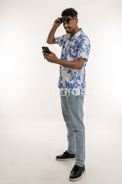Follow the latest social media news, see happy news, Saudi man using modern devices, portrait of a smiling dark-skinned young Arab Gulf Emirati man wearing casual clothes, removing sunglasses from his eyes and looking at the phone with gestures of pleasure, full body image, white background