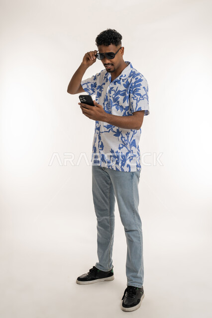 Browsing social networking sites online, the concept of elegance and interest in external appearance, portrait of a dark-skinned young Arab Gulf Emirati man wearing casual clothes and sunglasses holding a mobile phone in his hand, a Saudi man using modern devices, a full body image, white background
