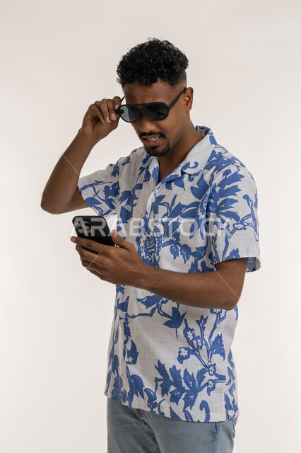 The concept of elegance and interest in external appearance, portrait of a dark-skinned young Arab Gulf Emirati man wearing casual clothes and sunglasses holding a mobile phone in his hand, a Saudi man using modern devices, browsing social networking sites on the Internet, white background