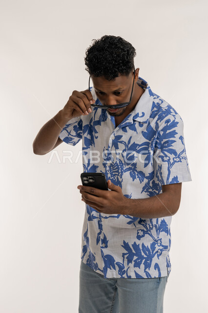 رؤية الاخبار المفاجئة، رجل سعودي ينظر الى الجوال بتعابير الدهشة والانبهار، بورتريه لشاب اسمر عربي خليجي إماراتي يرتدي الزي الكاجوال يقوم برفع النظارات الشمسية عن عينه والنظر الى الهاتف المحمول بإيماءات التعجب والإستغراب، خلفية بيضاء