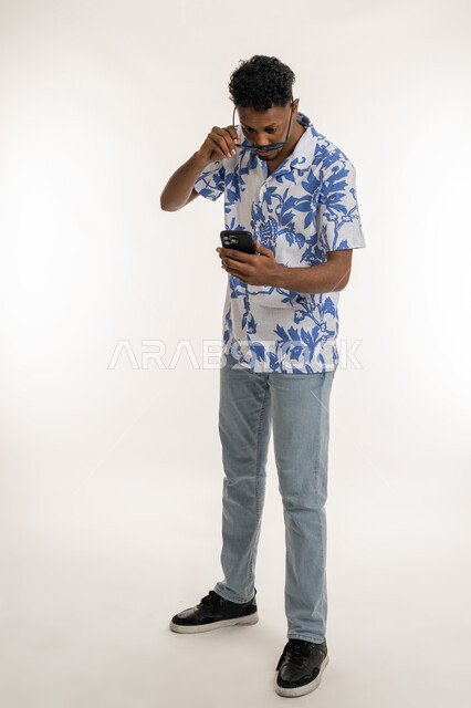 View of sudden news, a Saudi man looking at his mobile phone with expressions of astonishment and amazement, a portrait of a dark-skinned young Arab Gulf Emirati man wearing casual clothes, removing his sunglasses from his eyes and looking at his mobile phone with gestures of astonishment and surprise, full body image, white background
