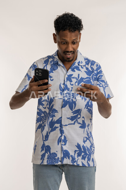 سهولة الشراء والدفع من خلال الهاتف المحمول، ايماءات الذهول والاندهاش، بورتريه لشاب اسمر عربي خليجي اماراتي يرتدي الزي الكاجوال يحمل بيده الجوال وبطاقة الإئتمان البنكية، شاب سعودي ينظر الى كرت الصراف بتعابير الدهشة, تسديد الفواتير بالبطاقات الإلكترونية، خلفية بيضاء