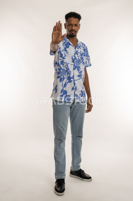 مفهوم الرفض والامتناع والانزعاج، طلب التوقف وعدم الاقتراب، بورتريه لشاب اسمر عربي خليجي اماراتي يرتدي الزي الكاجوال يمد كفه نحو الامام بتعابير النهي عن فعل شيء ما, رجل سعودي ينظر الى الكاميرا بايماءات الحزم والجدية, صورة الجسم بالكامل، خلفيه بيضاء