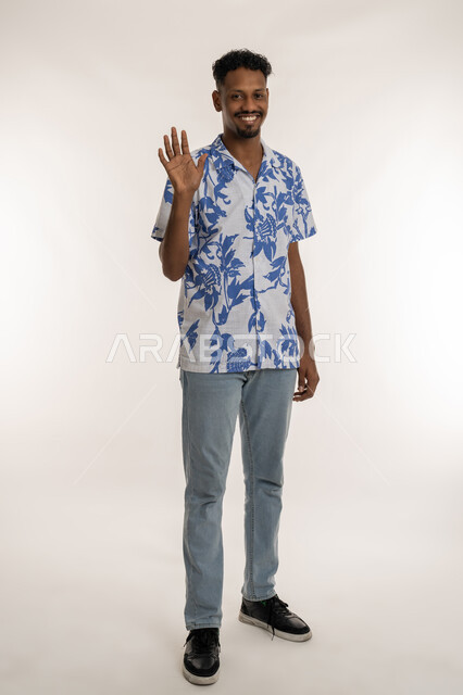مفهوم الاستقبال والترحيب، شاب سعودي ينظر الى لكاميرا بايماءات الفرح والسرور، بورتريه لرجل اسمر عربي خليجي اماراتي يرتدي الزي الكاجوال يرفع كف يده للاعلى بتعابير القاء التحية، التعارف و الصداقه وتكوين العلاقات، صورة الجسم بالكامل، خلفية بيضاء