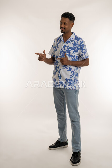 الإشارة و التوضيح وتحديد موقع معين, رجل سعودي يقوم بالدلالة على مكان عنصر، بورتريه لشاب اسمر عربي خليجي اماراتي مبتسم يرتدي الزي الكاجوال يشير بإصبعيه السبابة لشيء ما بايماءات الفرد والتصويب، صورة الجسم بالكامل، خلفية بيضاء