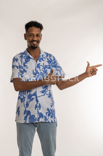 الدلالة على مكان عنصر، رجل سعودي يقوم بالإشارة و التوضيح وتحديد موقع معين، بورتريه لشاب اسمر عربي خليجي اماراتي يرتدي الزي الكاجوال ينظر الى الكاميرا و يشير بإصبعيه السبابة إلى شيء ما بايماءات الفرد والتصويب، خلفية بيضاء