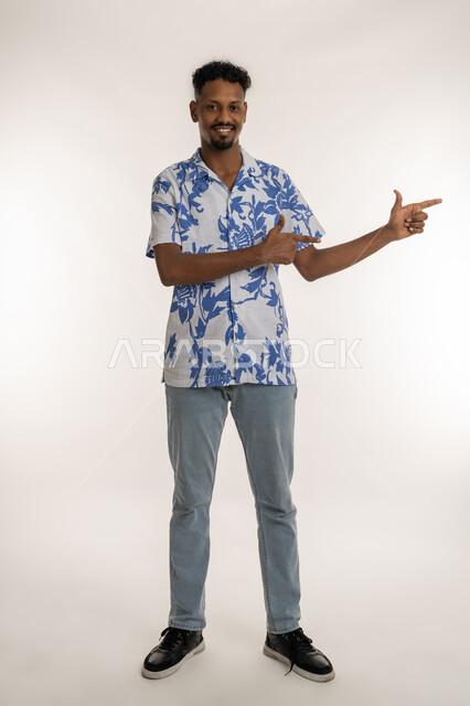 الإشارة و التوضيح وتحديد موقع معين, رجل سعوي يقوم بالدلالة على مكان عنصر، بورتريه لشاب اسمر عربي خليجي اماراتي يرتدي الزي الكاجوال ينظر الى الكاميرا و يشير بإصبعيه السبابة إلى شيء ما بايماءات الفرد والتصويب، صورة الجسم بالطول الكامل، خلفية بيضاء
