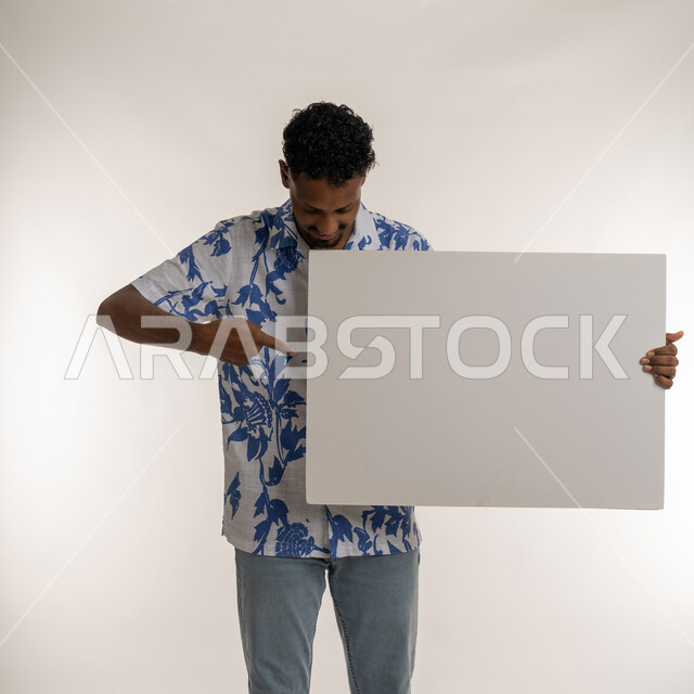 لافتة إعلانية فارغة، استخدام لوحات توضيحية، مفهوم الإعلان والتسويق، شاب سعودي يحمل موك اب أبيض مستطيل، بورتريه لرجل عربي خليجي اماراتي مبتسم يرتدي الزي الكاجوال يشير باصبع السبابة للوحه بيضاء، صورة لكامل الجسم، خلفية بيضاء
