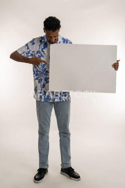 استخدام لوحات توضيحية، لافتة إعلانية فارغة، مفهوم الإعلان والتسويق، شاب سعودي يحمل موك اب أبيض مستطيل، بورتريه لرجل عربي خليجي اماراتي مبتسم يرتدي الزي الكاجوال يشير باصبع السبابة للوحه بيضاء، صورة لكامل الجسم، خلفية بيضاء