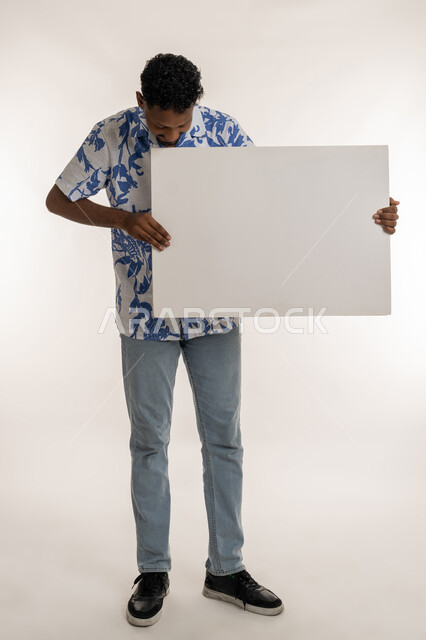 موك اب أبيض مستطيل، مفهوم الإعلان والتسويق، بورتريه لرجل عربي خليجي اماراتي مبتسم يرتدي الزي الكاجوال يحمل بيده لوحه بيضاء، شاب سعودي يمسك لافتة إعلانية فارغة ،استخدام لوحات توضيحية، صورة لكامل الجسم، خلفية بيضاء
