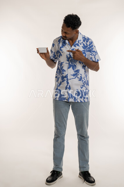 Empty rectangular card, illustration of the importance of personal identity, a young Saudi man holding a white mockup, portrait of a smiling Arab Gulf Emirati man in casual wear pointing his index finger at a bank card, full body length, white background