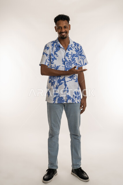 حركات يدين تدل على هذا المكان"هنا"، تحديد موقع شيء معين، رجل سعودي يشير بيده باتجاه اليسار، بورتريه لرجل عربي خليجي اماراتي يرتدي الزي الكاجوال يشير بيده لجهة اليسار ينظر للكاميرا بايماءات البشاشة، مفهوم الأناقة والاهتمام بالمظهر الخارجي، صورة لكامل الجسم، خلفيه بيضاء