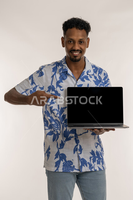 Blank black screen display, integrating advanced technology into practical life, Saudi man using modern technology, portrait of a smiling dark-skinned young Arab Gulf Emirati man wearing casual clothes pointing his finger at his laptop, white background