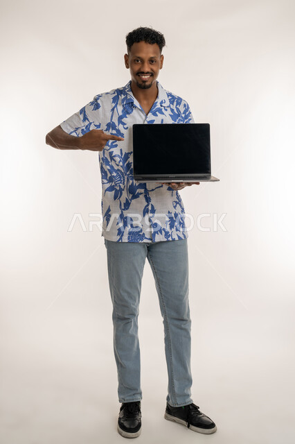 Integrating advanced technology with practical life, Saudi man using modern technology, portrait of a smiling dark-skinned young Arab Gulf Emirati man in casual wear pointing his finger at laptop, blank black screen display, full body image, white background
