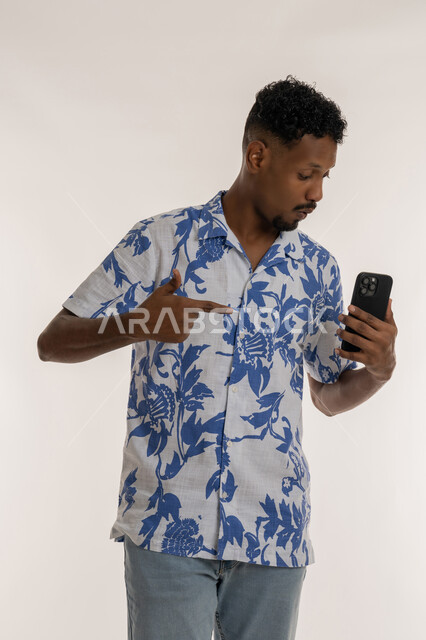 Benefiting from mobile services and applications, integrating technology and modern devices into daily life, a Saudi young man chatting and communicating with family and friends, portrait of a smiling Emirati Gulf Arab man wearing casual clothes pointing his index finger at a mobile phone, white background