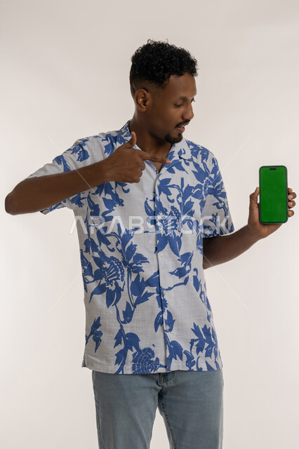 دمج التكنولوجيا والاجهزة الحديثة في الحياة اليومية، تقديم خدمات وتطبيقات الجوال، بورتريه لرجل عربي خليجي اماراتي مبتسم يرتدي الزي لكاجوال يشير باصبع السبابة للهاتف محمول، شاب سعودي يقوم بعرض شاشة خضراء فارغة، خلفية بيضاء