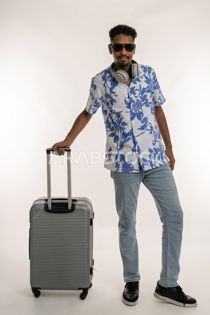 قضاء رحلة صيفية ممتعة, رجل سعودي ينظر إلى الكاميرا بتعابير الفرح والسرور، بورتريه لشاب عربي خليجي إماراتي مبتسم يرتدي الزي الكاجوال والنظارة الشمسية ويضع الهيدفون حول عنقه يمسك بيده حقيبة السفر, جولات ترفيهية ممتعة في الإجازة، صورة لكامل الجسم، خلفية بيضاء