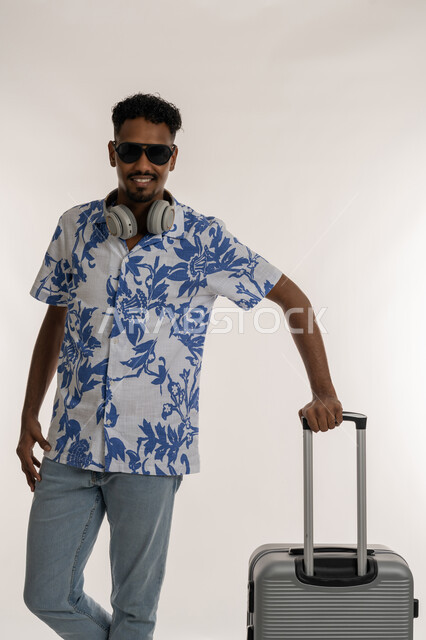 جولات ترفيهية ممتعة في الإجازة، الاستعداد لقضاء رحلة صيفية ممتعة, رجل سعودي ينظر إلى الكاميرا بتعابير الفرح والسرور، بورتريه لشاب عربي خليجي إماراتي مبتسم يرتدي الزي الكاجوال والنظارة الشمسية ويضع الهيدفون حول عنقه يمسك بيده حقيبة السفر, خلفية بيضاء