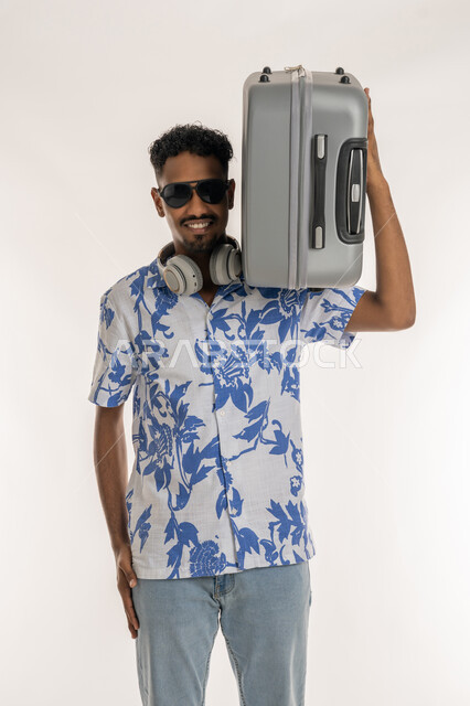 النظر الى الكاميرا بتعابير السرور، الاستعداد لقضاء رحلة صيفية ممتعة, بورتريه لشاب عربي خليجي إماراتي مبتسم يرتدي الزي الكاجوال والنظارة الشمسية يضع الهيدفون حول عنقه يحمل حقيبة السفرعلى كتفه، رجل سعودي يقوم بجولة ترفيهية في الإجازة، خلفية بيضاء