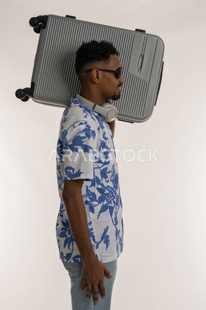 قضاء رحلة صيفية ممتعة, بورتريه من الجانب لشاب عربي خليجي إماراتي مبتسم يرتدي الزي الكاجوال والنظارة الشمسية يضع الهيدفون حول عنقه يحمل حقيبة السفرعلى كتفه، رجل سعودي يقوم بجولة ترفيهية في الإجازة، خلفية بيضاء