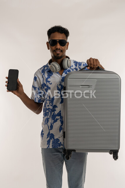 جولات ترفيهية في الإجازة، الاستعداد لقضاء رحلة صيفية ممتعة, بورتريه لشاب عربي خليجي إماراتي مبتسم يرتدي الزي الكاجوال والنظارة الشمسية ويضع الهيدفون حول عنقه يحمل حقيبة السفرعلى كتفه، رجل سعودي يمسك بيده هاتف محمول بشاشة سوداء، خلفية بيضاء