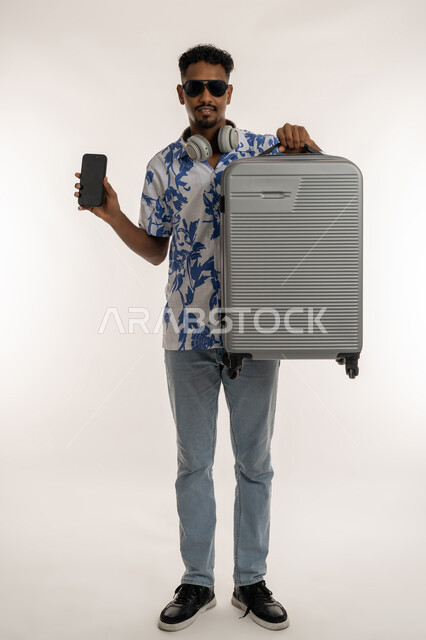 الاستعداد لقضاء رحلة صيفية ممتعة, جولات ترفيهية في الإجازة، بورتريه لشاب عربي خليجي إماراتي مبتسم يرتدي الزي الكاجوال والنظارة الشمسية ويضع الهيدفون حول عنقه يمسك حقيبة السفر، رجل سعودي يحمل بيده هاتف محمول بشاشة سوداء, صورة لكامل الجسم، خلفية بيضاء