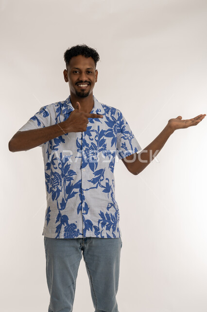 حمل عنصر هولوجرامي، تحديد موقع شيء ما، بورتريه لشاب اسمر عربي خليجي اماراتي مبتسم يرتدي الزي الكاجوال يشير الى كفه بايماءات السرور والسعادة، مفهوم التقديم والعرض، رجل سعودي ينظر الى الكاميرا بتعابير البهجة، خلفية بيضاء